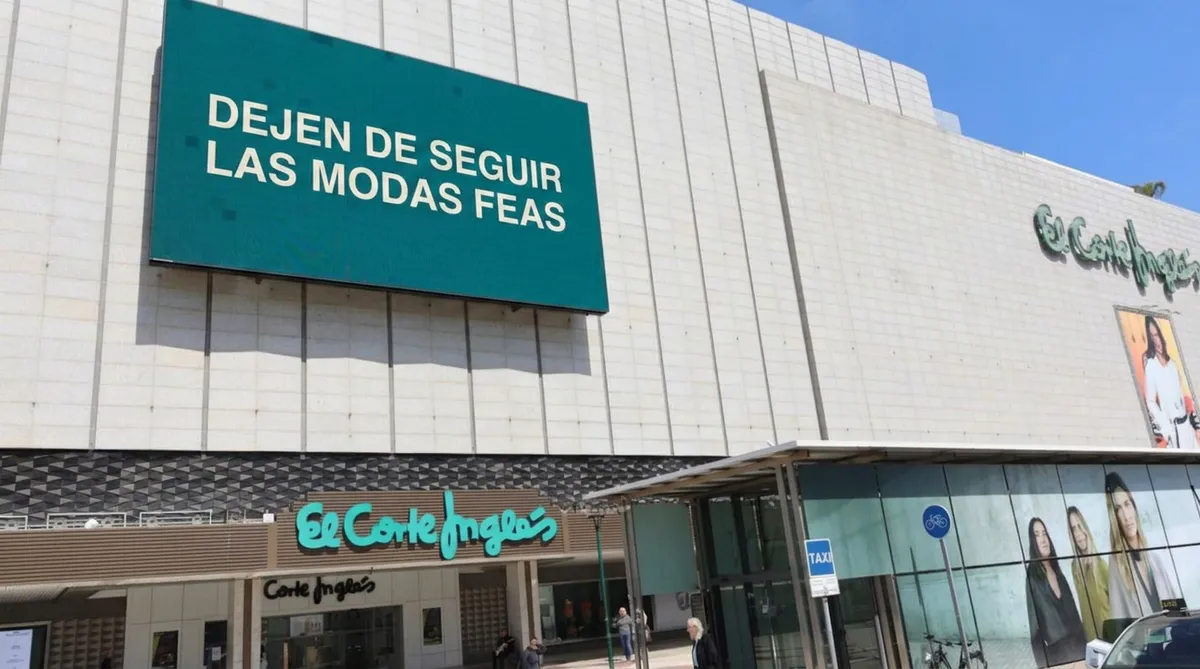 The large LED screen above the El Corte Inglés entrance in Málaga displaying "DEJEN DE SEGUIR LAS MODAS FEAS" in white text on a teal background.