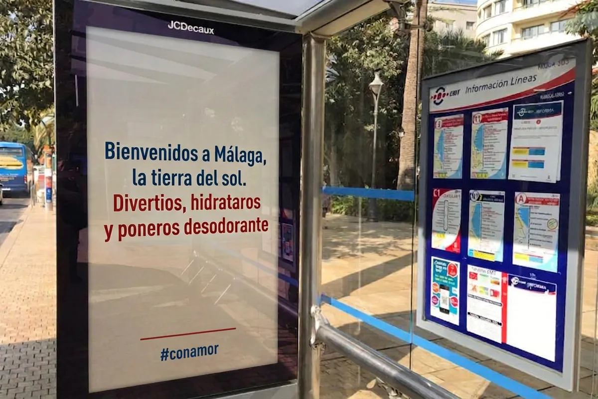 A sign at Málaga bus stops reading "Welcome to Málaga, land of the sun — have fun, stay hydrated and put on deodorant."