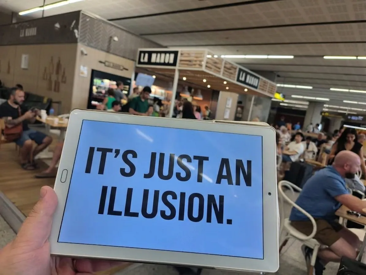A tablet held by a driver at the Málaga airport arrivals area, displaying "IT'S JUST AN ILLUSION" instead of a passenger's name.