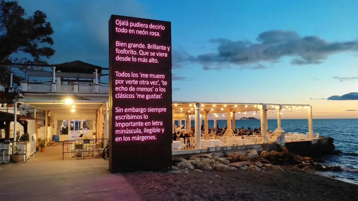 A pink neon-style sign on a dark wall, with a long message in Spanish: "Ojalá pudiera decirlo todo en neón rosa. Bien grande, brillante y fosforito. Que se viera desde lo más alto. Todos los 'me muero por verte otra vez', 'te echo de menos' o los clásicos 'me gustas'. Sin embargo siempre escribimos lo importante en letra minúscula, ilegible y en los márgenes."